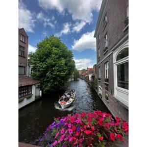 Home Tourists enjoy a scenic boat ride along a historic canal in Bruges, surrounded by flowers and architecture.